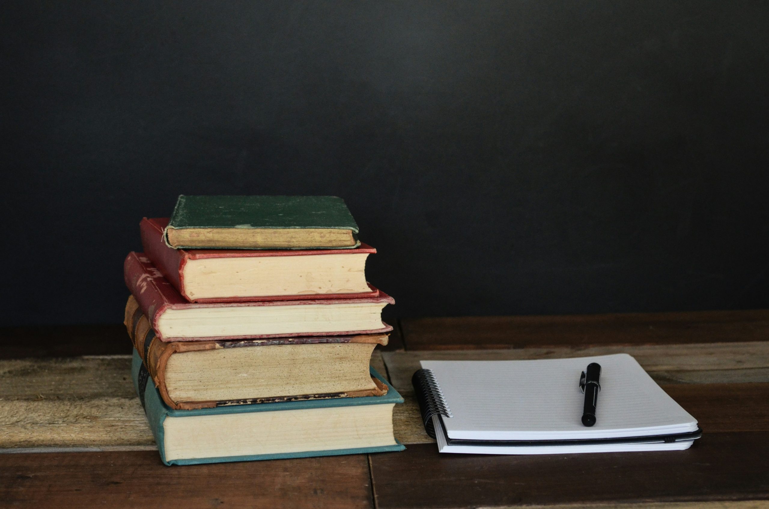 A stack of old books sits beside an open notebook and a pen, suggesting a study or writing scene.