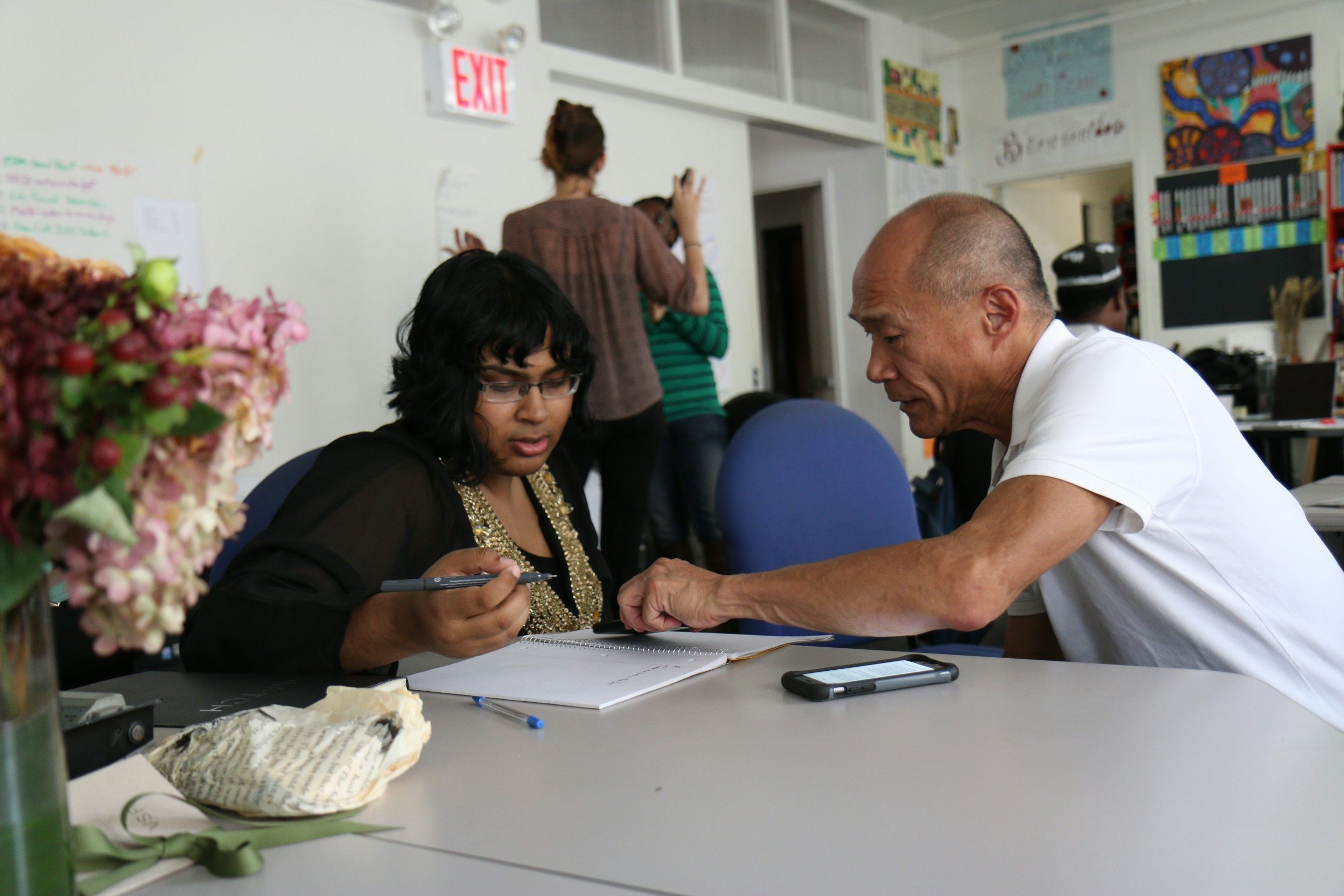 A woman writes at a table while a man sits across from her, likely in a classroom or study.