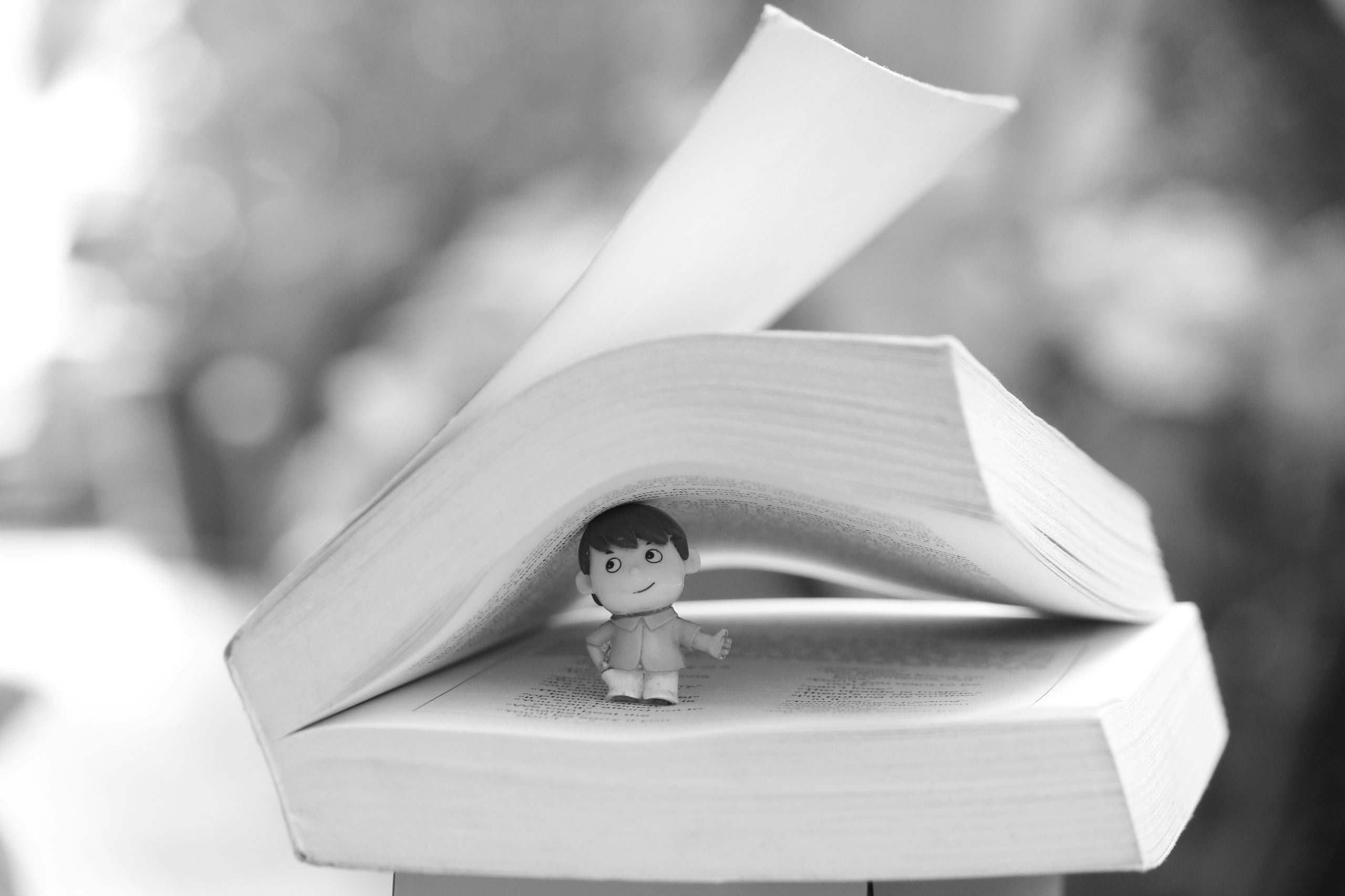 A grayscale stock photo of a miniature figure sitting inside the pages of an open book.