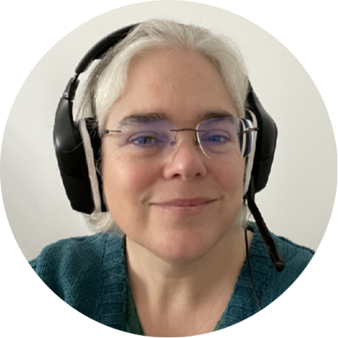 A middle-aged white woman with grey hair, glasses, and headphones, smiling in a green jumper.