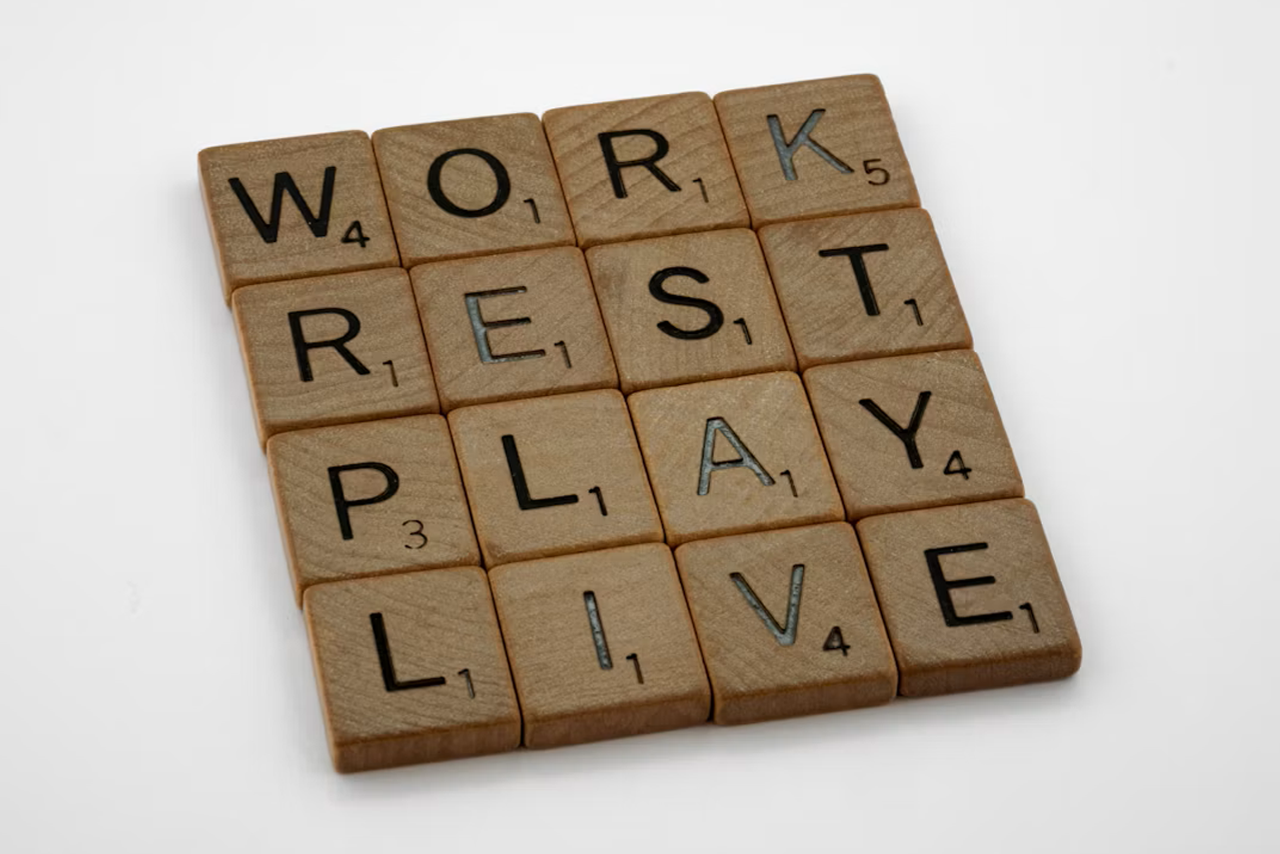 Wooden letter tiles arranged in a four-by-four grid spelling out the words "WORK", "REST", "PLAY", and "LIVE" on a plain white background.